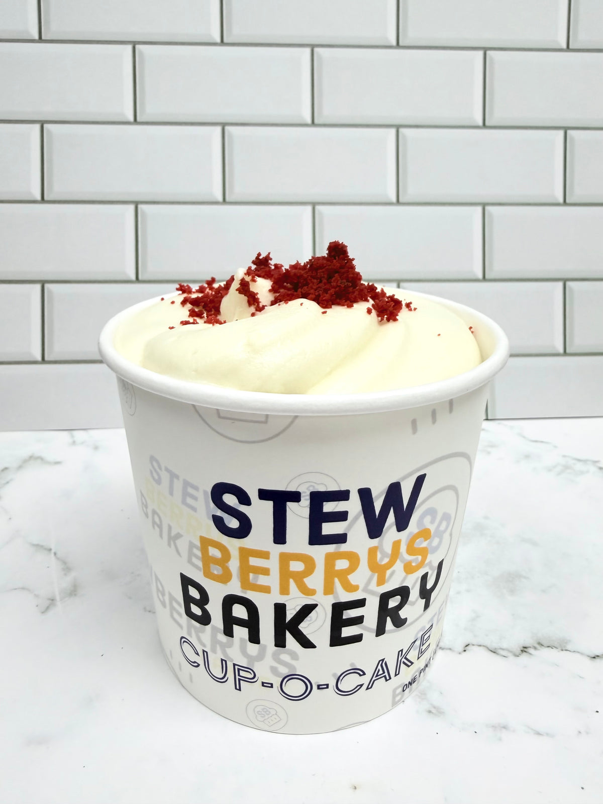 A white paper cup of cream cheese frosting of gluten free red velvet cake with red cake crumbs  branded 'StewBerry's Bakery', set on a white marble surface against a white subway tile background.