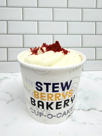 A white paper cup of cream cheese frosting of gluten free red velvet cake with red cake crumbs  branded 'StewBerry's Bakery', set on a white marble surface against a white subway tile background.