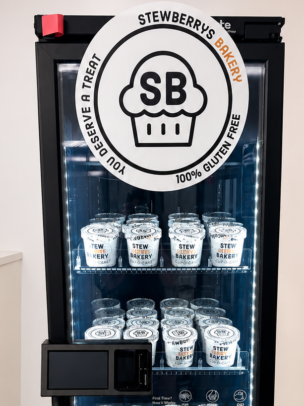 A Stewberry's Bakery branded glass-door vending machine or cooler filled with small white cup containers of gluten free cake, with a large circular SB logo decal on the door.